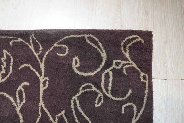 Dark brown high-pile rug with a beige scrolling vine pattern rests on light wood planks.