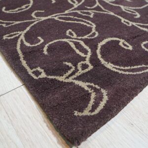 Dark plum rug with thick, beige scrollwork pattern rests on pale blonde wood flooring.
