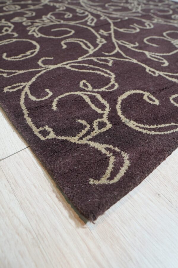 Dark plum rug with thick, beige scrollwork pattern rests on pale blonde wood flooring.