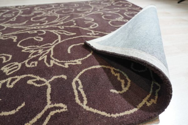 Dark brown medium-pile rug with beige scrolling pattern, rolled up on light wood floor.