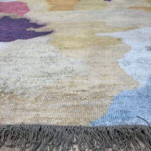 Close-up of an abstract rug featuring purple, pink, blue, and yellow shapes on light wood flooring with a gray fringe.