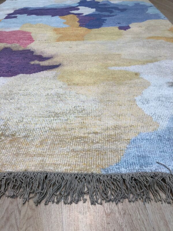 Close-up of an abstract rug featuring purple, pink, blue, and yellow shapes on light wood flooring with a gray fringe.