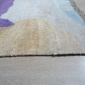 Detail of a textured, fringed rug with abstract purple, cream, and blue colors on light hardwood.