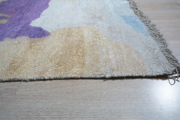 Detail of a textured, fringed rug with abstract purple, cream, and blue colors on light hardwood.