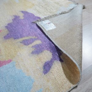 Abstract pink, purple, and blue rug shown partially rolled back, exposing woven backing on light wood flooring.