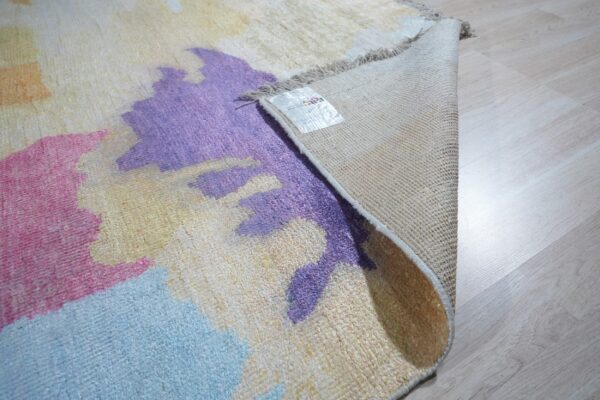 Abstract pink, purple, and blue rug shown partially rolled back, exposing woven backing on light wood flooring.