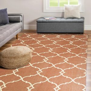 A rust orange flatweave rug with a cream quatrefoil pattern sits on wood floors near gray furniture.