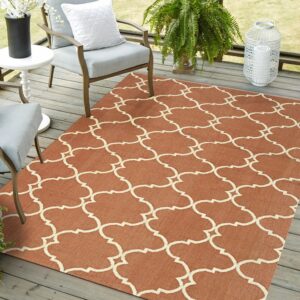 A terracotta rug with a cream quatrefoil lattice pattern sits on a gray wood deck with patio chairs and plants.