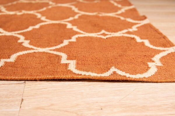 Low-angle view of a flat woven orange and cream rug featuring a trellis pattern on blonde wood planks.