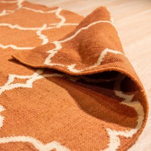 Rust orange flatweave rug with off-white geometric pattern is rolled up on light wood plank flooring.