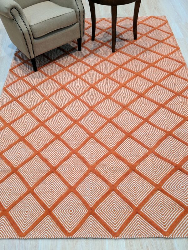 Refined luxurious yellow handmade wool rug 3 Textured orange and white rug with concentric diamonds placed beneath a beige armchair and dark wooden table legs.