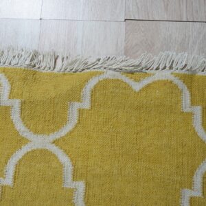 Yellow flatweave rug featuring a white quatrefoil pattern and twisted fringe on pale wood planks.