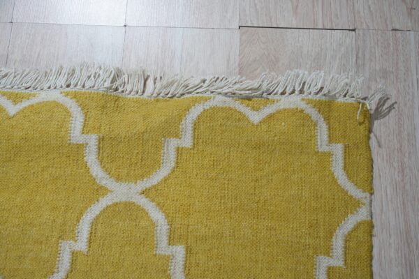 Yellow flatweave rug featuring a white quatrefoil pattern and twisted fringe on pale wood planks.