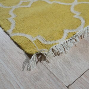 Yellow rug with white trellis pattern and fringe detail rests on pale wood planks.