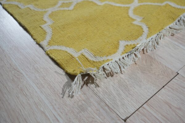 Yellow rug with white trellis pattern and fringe detail rests on pale wood planks.