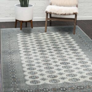 Cream rug with blue and tan geometric medallions lies on dark floor next to a white furry chair.