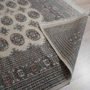 Cream rug with blue and peach geometric patterns partially rolled back on light wood flooring.