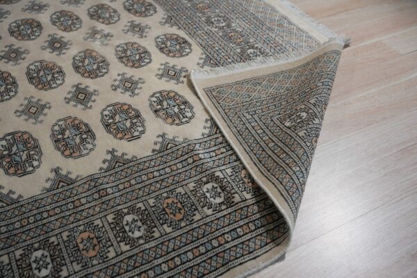 Cream rug with blue and peach geometric patterns partially rolled back on light wood flooring.