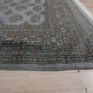 Low-pile gray rug featuring yellow, black, and teal traditional patterns and fringe on light laminate flooring.