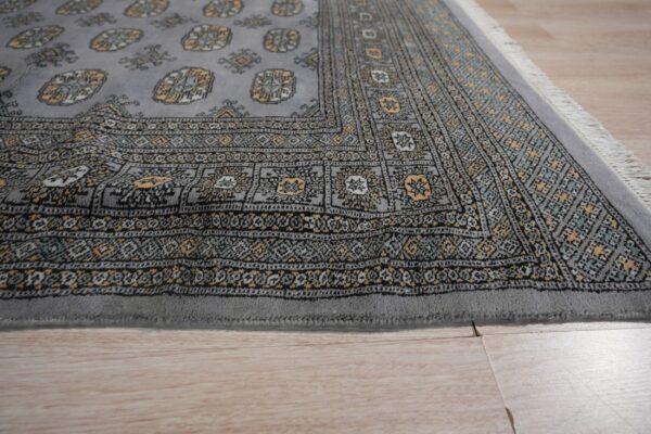 Low-pile gray rug featuring yellow, black, and teal traditional patterns and fringe on light laminate flooring.
