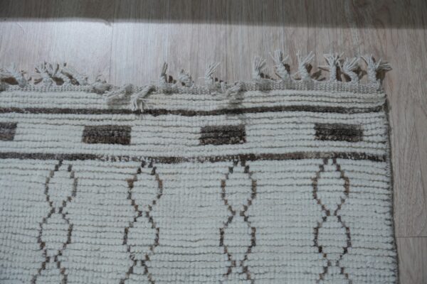 Textured cream rug featuring brown striped and diamond patterns, visible fringe, laid on light wood planks.