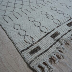 Textured off-white rug with dark brown lattice patterns and thick fringe rests on light wood floorboards.