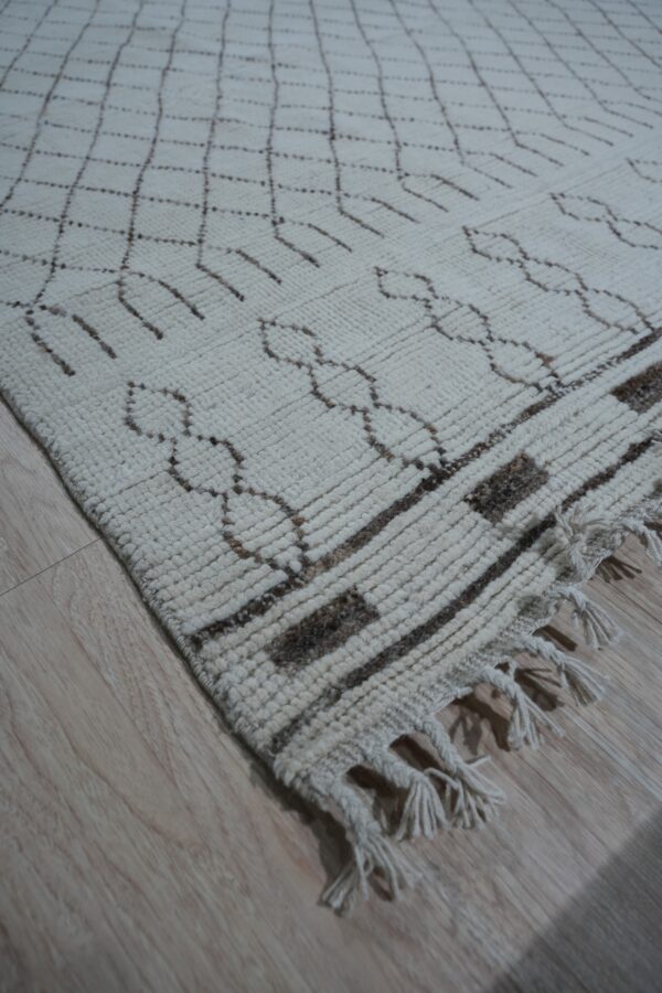 Textured off-white rug with dark brown lattice patterns and thick fringe rests on light wood floorboards.