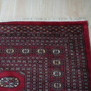 Red traditional rug with geometric patterns and fringe lies on light blonde wood flooring.