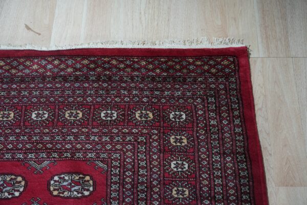 Red traditional rug with geometric patterns and fringe lies on light blonde wood flooring.