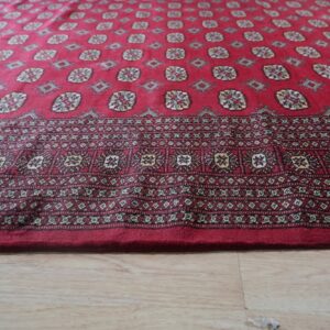 Deep red rug with complex geometric patterns and a wide border on light wood plank flooring.
