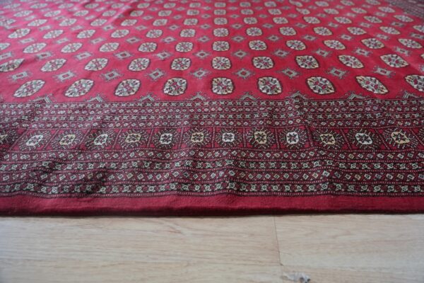 Deep red rug with complex geometric patterns and a wide border on light wood plank flooring.