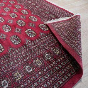 A red patterned rug featuring tan and white geometric motifs is folded on light wood flooring.