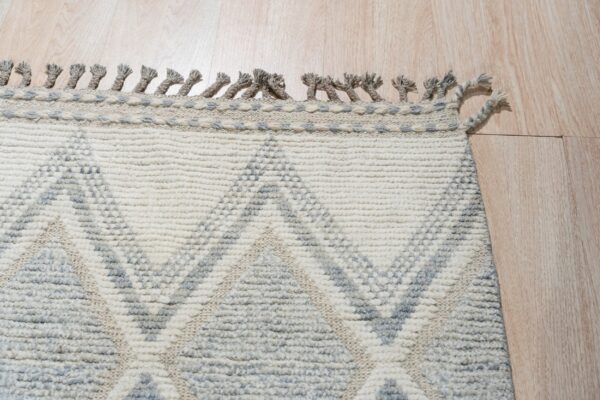 Textured cream and gray geometric pattern rug with braided tassels resting on a light wooden floor.