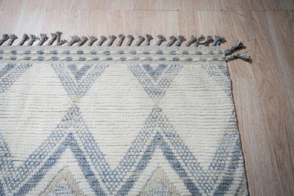 A nubby cream rug with blue geometric designs and braided fringe rests on light plank flooring.