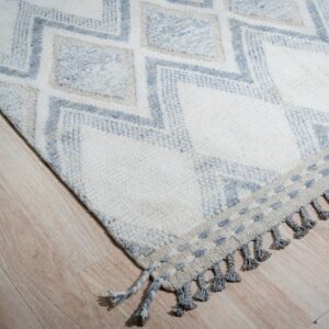 Textured ivory rug with blue diamond pattern, decorative fringe, and gray tassels on light wood planks.