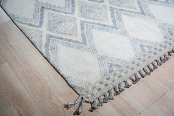 Textured ivory rug with blue diamond pattern, decorative fringe, and gray tassels on light wood planks.