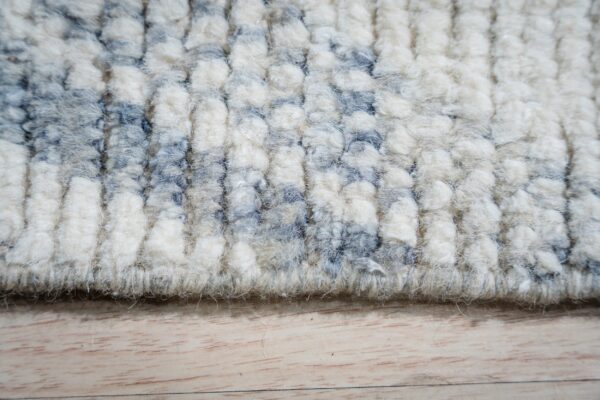 High-pile rug texture featuring thick rows of cream and dusty blue abstract patterns on light wood flooring.