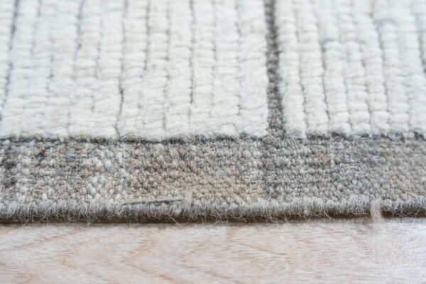Highly textured cream and gray striped rug edge transitioning to a dense low pile border on light wood floor.