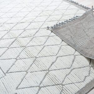 High-pile cream and gray diamond rug folded to show its striped gray woven backing and tassels on wood flooring.