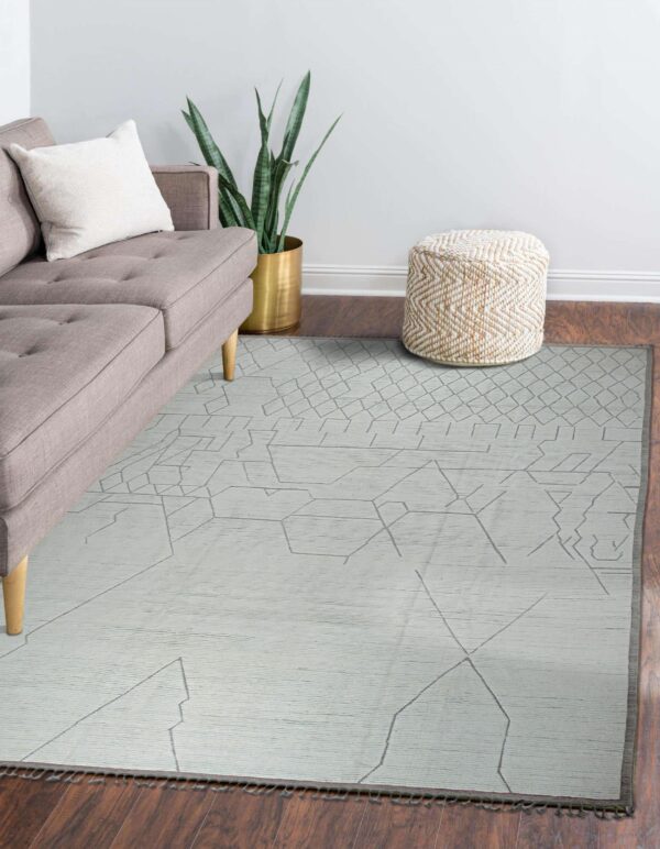 Pale low-pile rug with abstract geometric line patterns displayed in a room with a gray sofa and woven pouf.