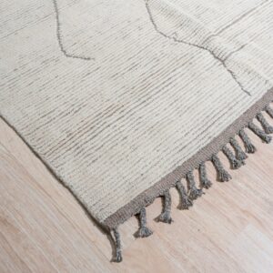 High-pile beige rug with geometric pattern and thick gray tassels lying on light wood planks.