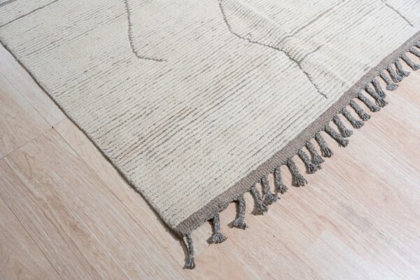 High-pile beige rug with geometric pattern and thick gray tassels lying on light wood planks.