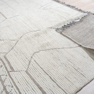 A textured cream rug with a taupe abstract pattern is partially rolled, showing the striped backing on light wood floor.
