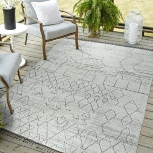 A low-pile gray geometric rug with dark abstract patterns and tassels sits beneath patio chairs on wood decking.