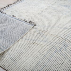 High-pile cream ribbed rug with subtle blue pattern, folded to show the striped flat-woven backing.
