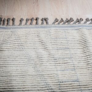 Textured cream rug with gray tassels and thin blue stripes lies on light wood flooring under directional lighting.