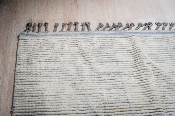 Textured cream rug with gray tassels and thin blue stripes lies on light wood flooring under directional lighting.