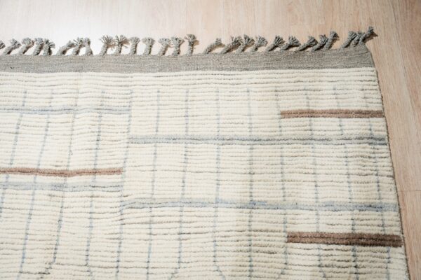 High-pile ivory rug featuring gray tassels, a subtle grid pattern, and horizontal brown accents on wood floor.