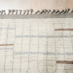High pile ivory rug featuring a grey grid, brown stripes, braided tassels, and light wood flooring.