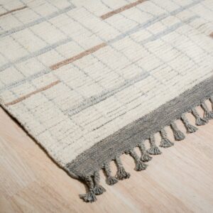Shaggy cream rug with gray grid and taupe stripes, bordered by dark gray fringe, on light wood flooring.